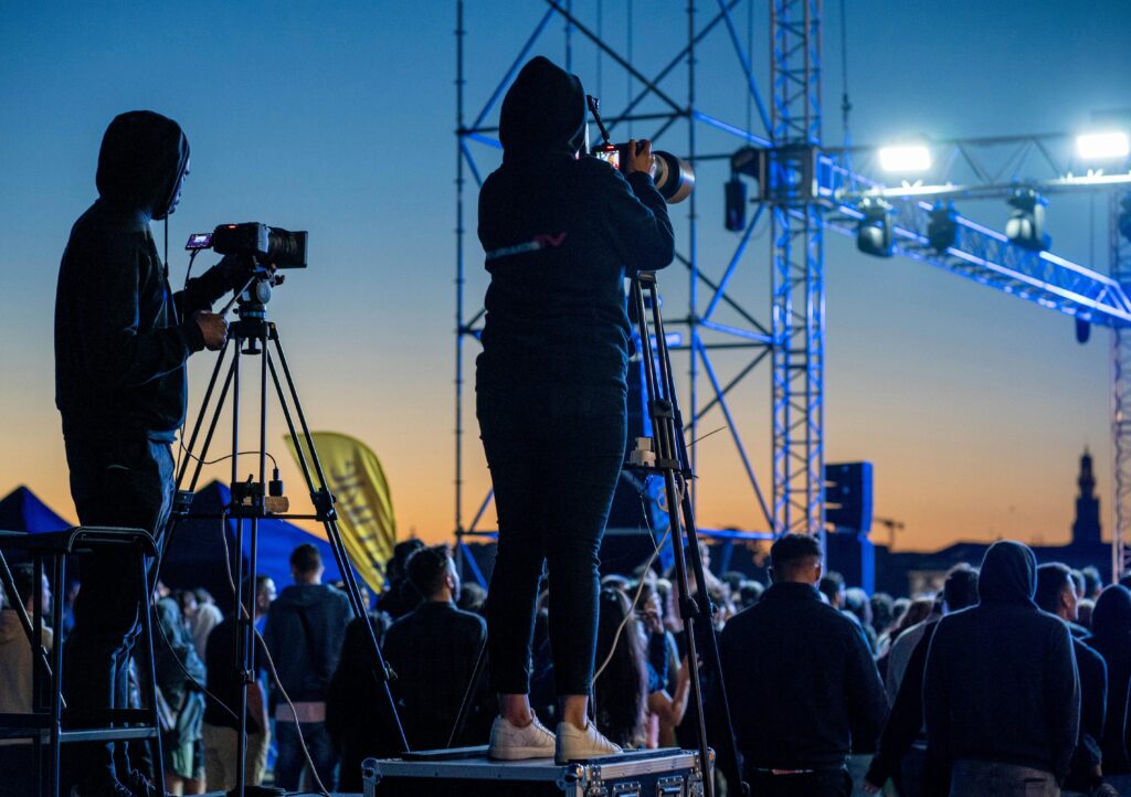 Photographers filming a vibrant outdoor event during a stunning sunset with an enthusiastic crowd.
